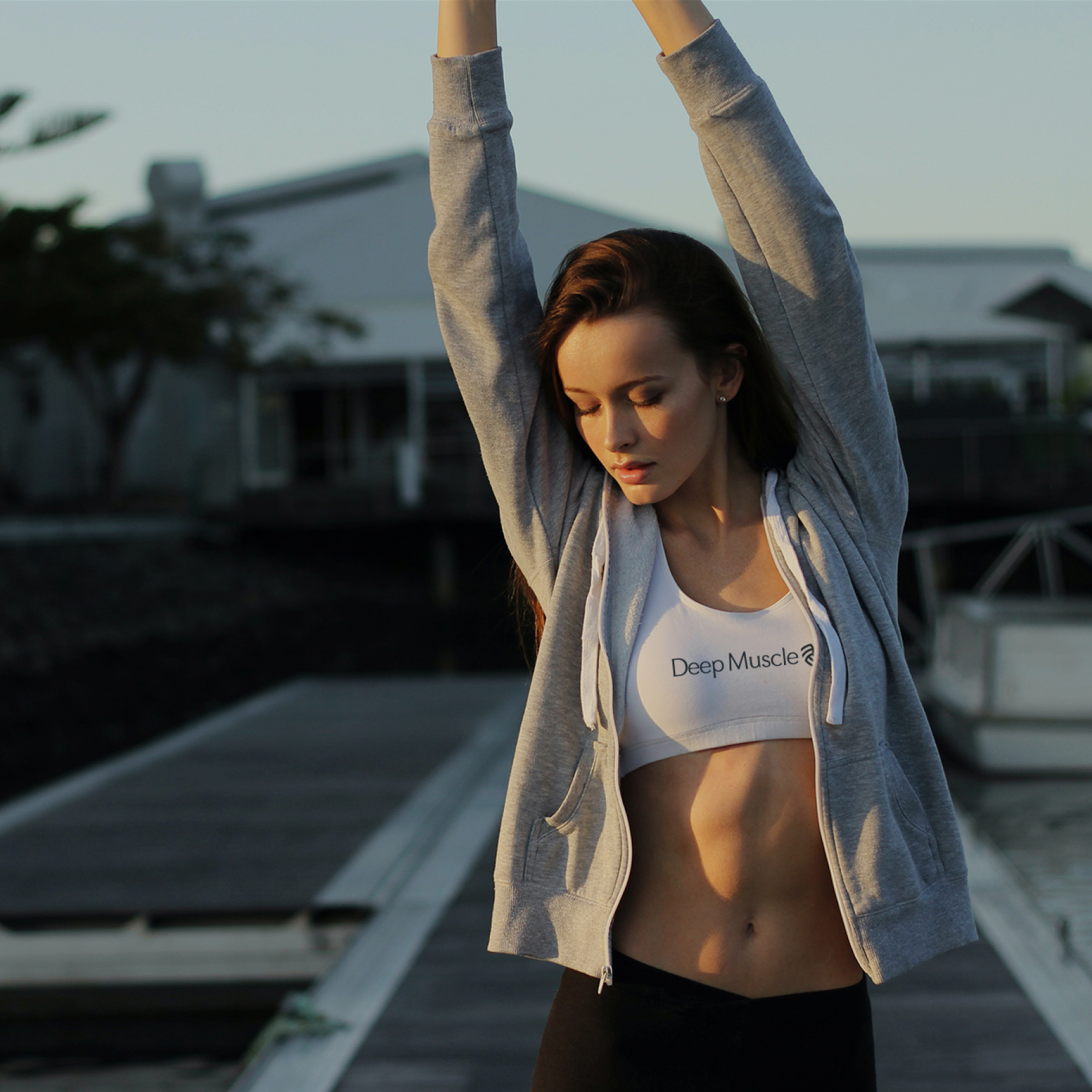 Woman in sports bra and hoodie stretching with arms raised by a dock at sunset.