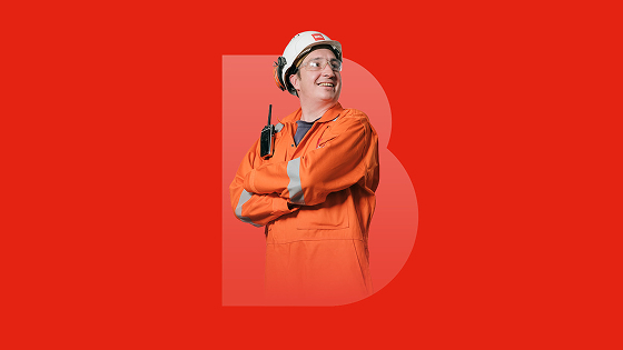 Man in orange safety coverall and white helmet with a walkie-talkie smiling with arms crossed on red background with large transparent letter B.