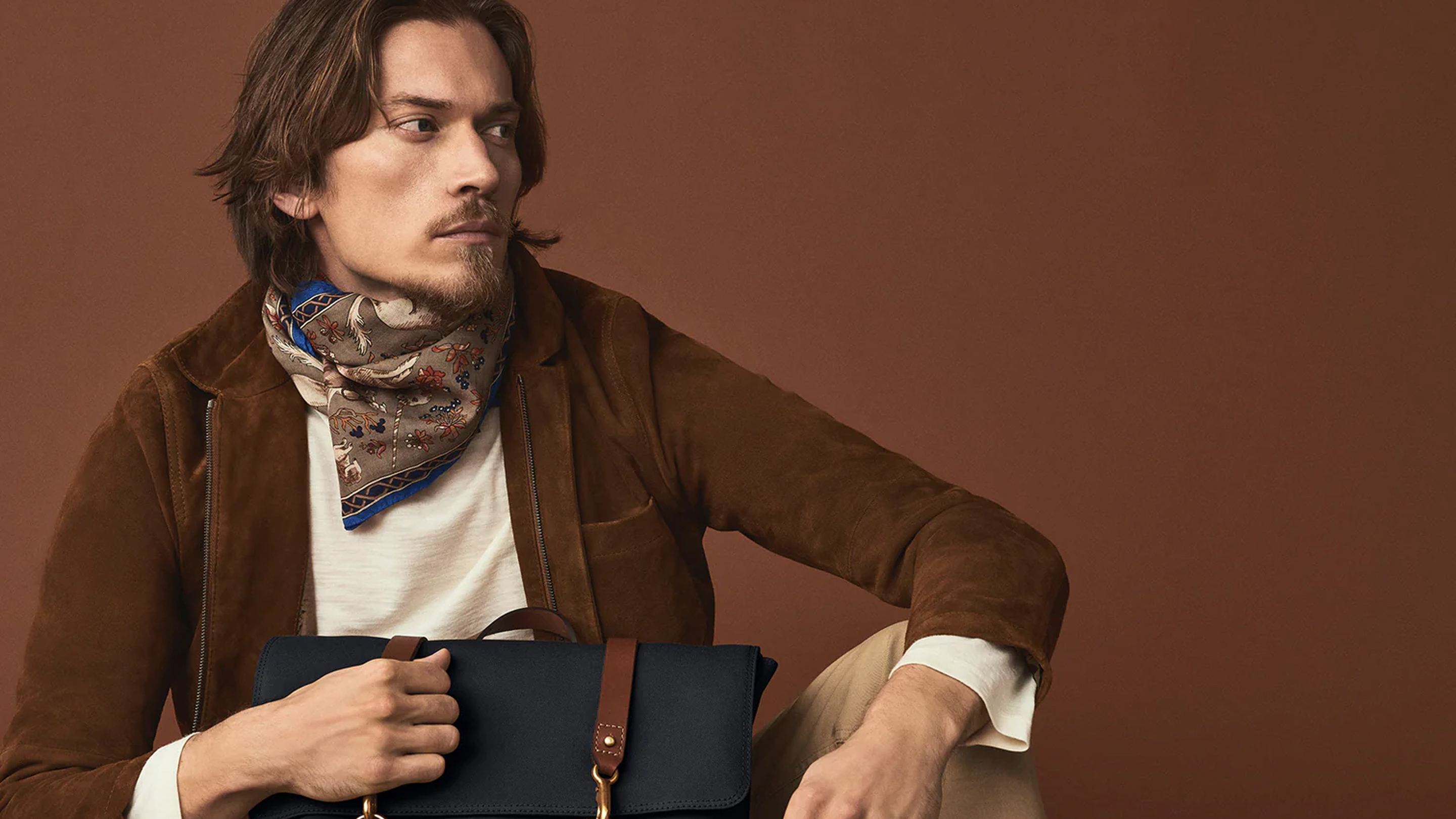 Stylish man with a brown jacket and patterned scarf holding a black leather briefcase against a brown background.