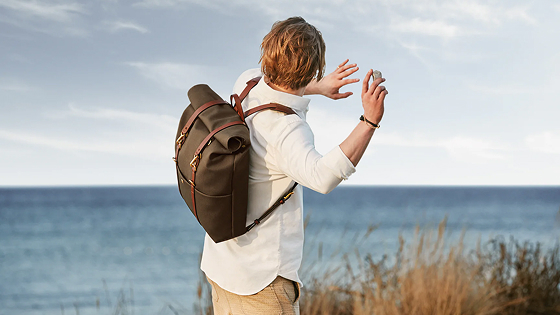 Person wearing a backpack throwing a small stone or object near a sea shore with dry grass in the foreground.