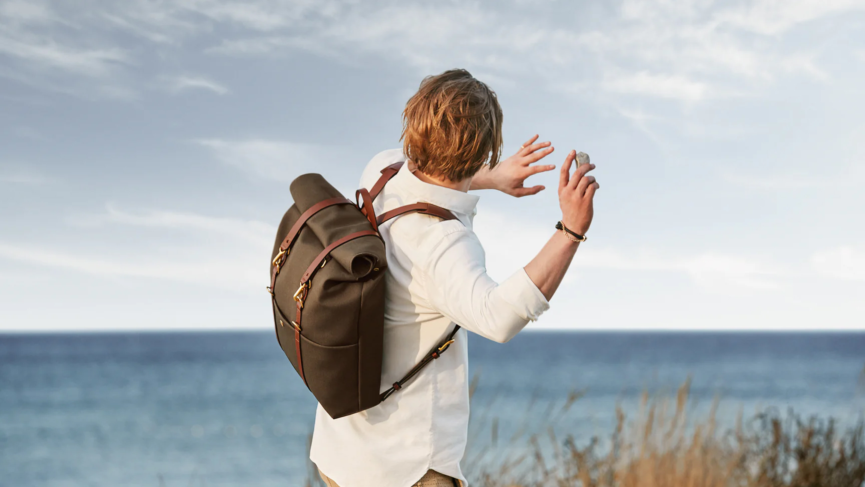 Person wearing a white shirt and a large dark backpack, throwing a stone toward the ocean under a partly cloudy sky.