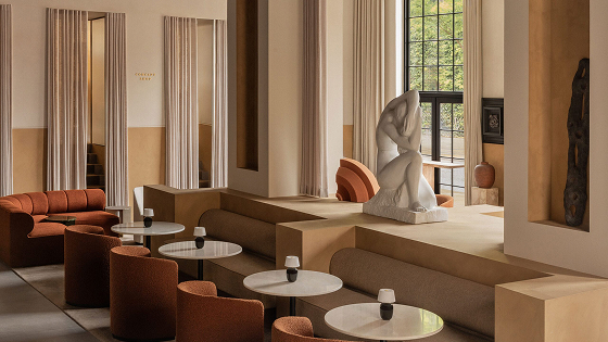Modern café interior with beige walls, rust-colored chairs, round marble tables, and a large white sculpture near a window.