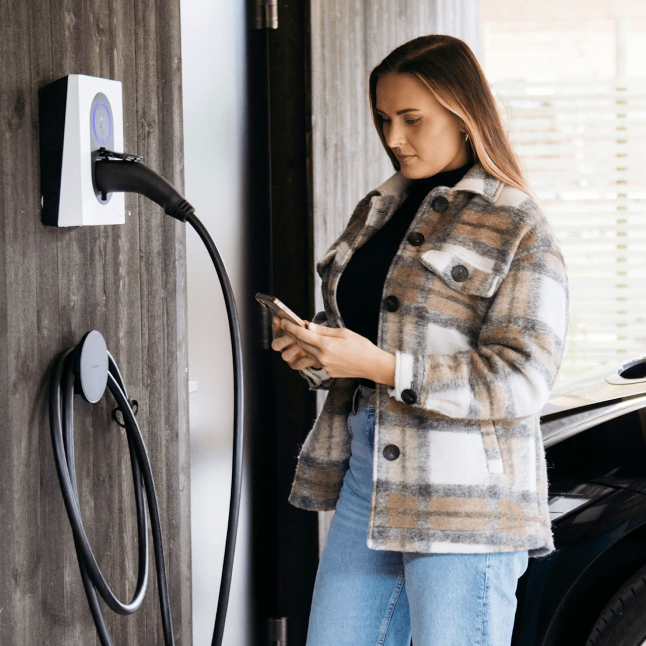Woman standing next to an electric vehicle charging station, holding a smartphone with a black car partially visible behind her.