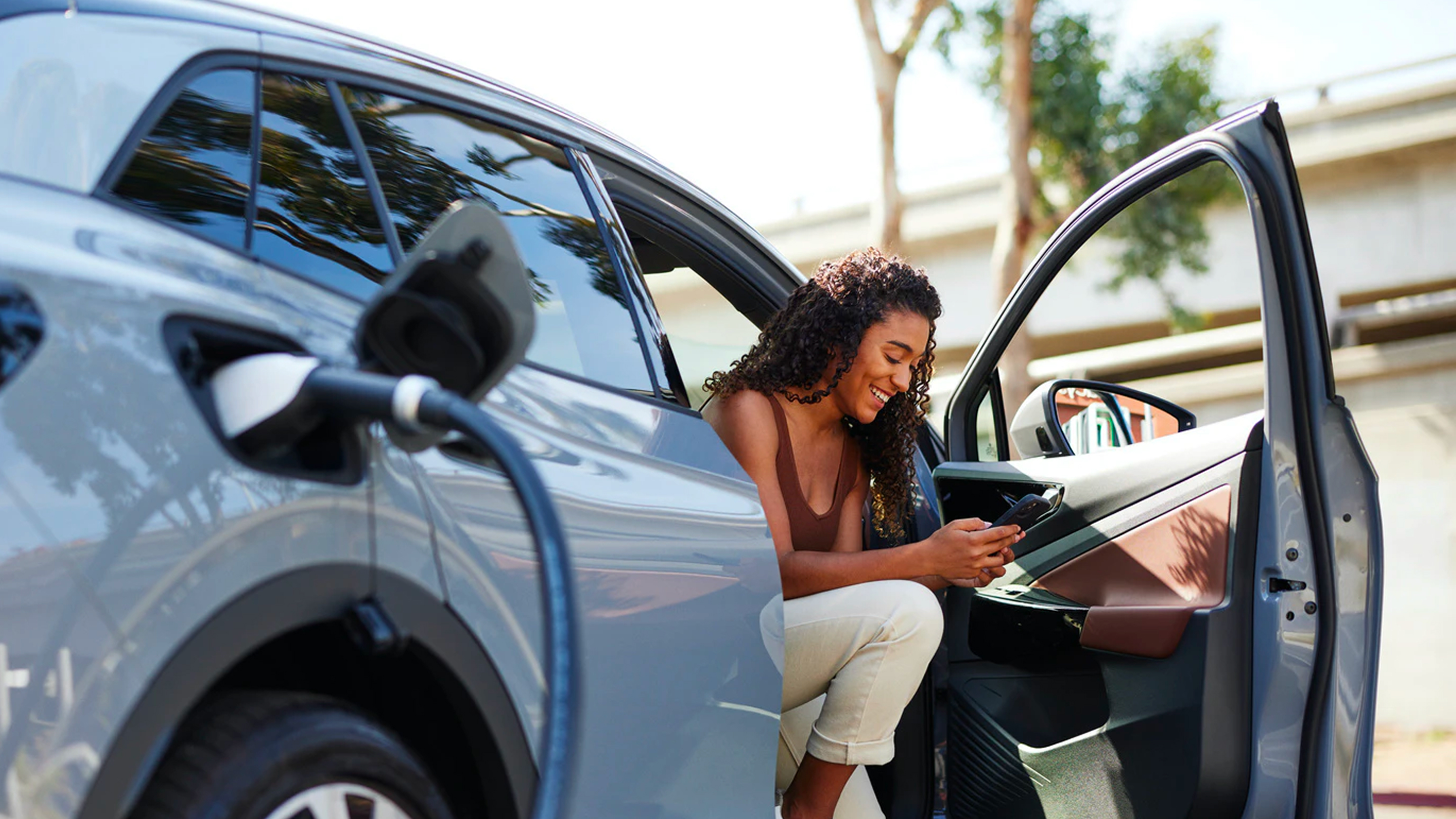 Woman sitting in an electric car with the door open, charging the vehicle and using a smartphone.