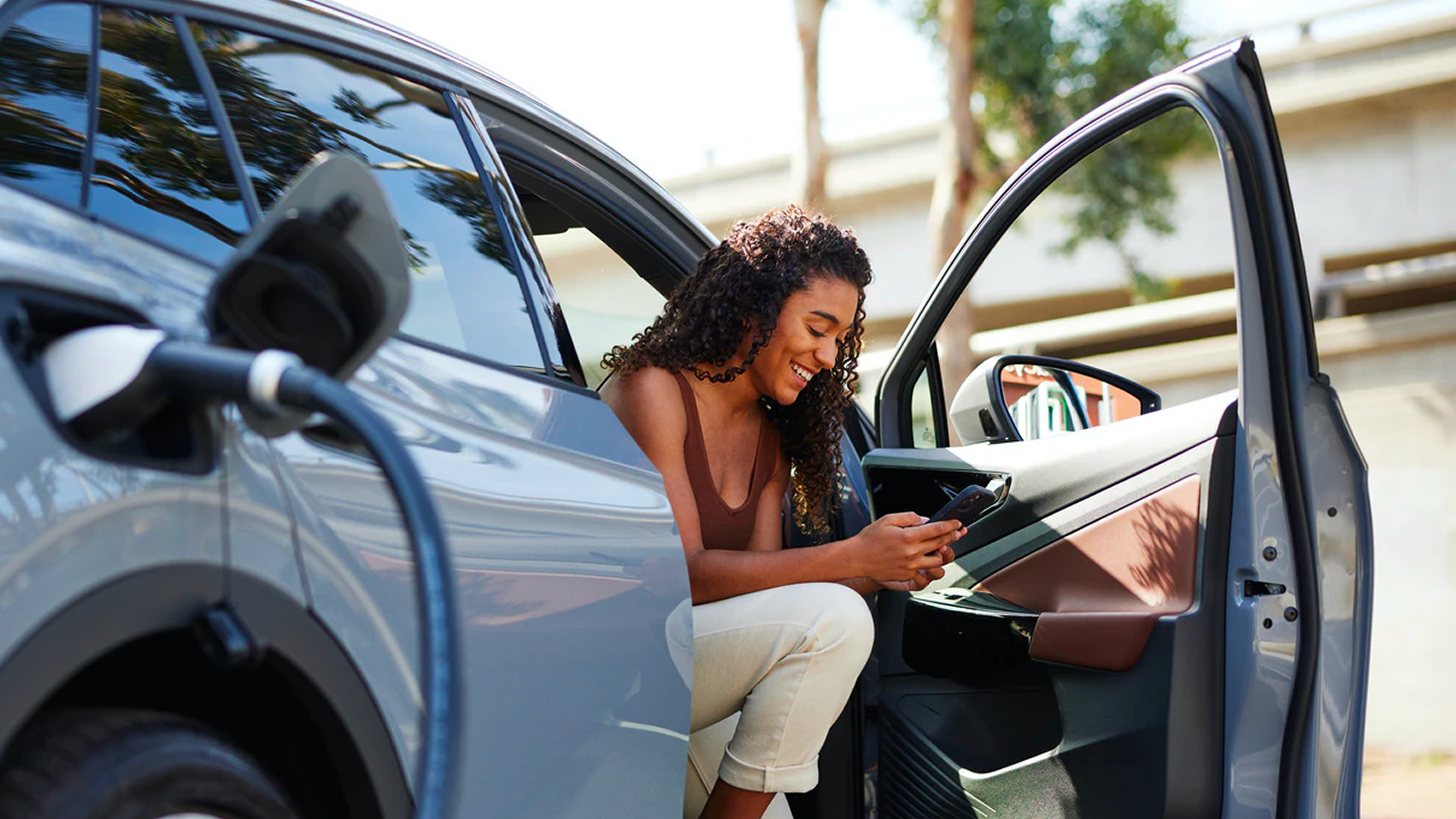Smiling woman sitting in an electric car with the door open, holding a phone while the car is charging.