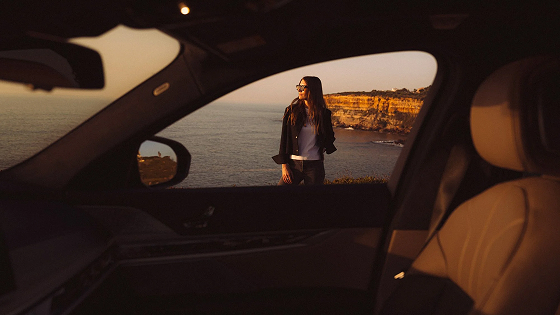 Woman with sunglasses standing by the ocean cliffs, viewed through a car window.